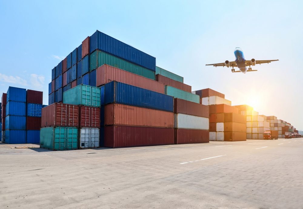 Stacked shipping containers at a port terminal with a cargo plane flying overhead at sunset
